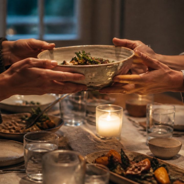 Hands passing a stoneware serving bowl across a linen-covered dinner table