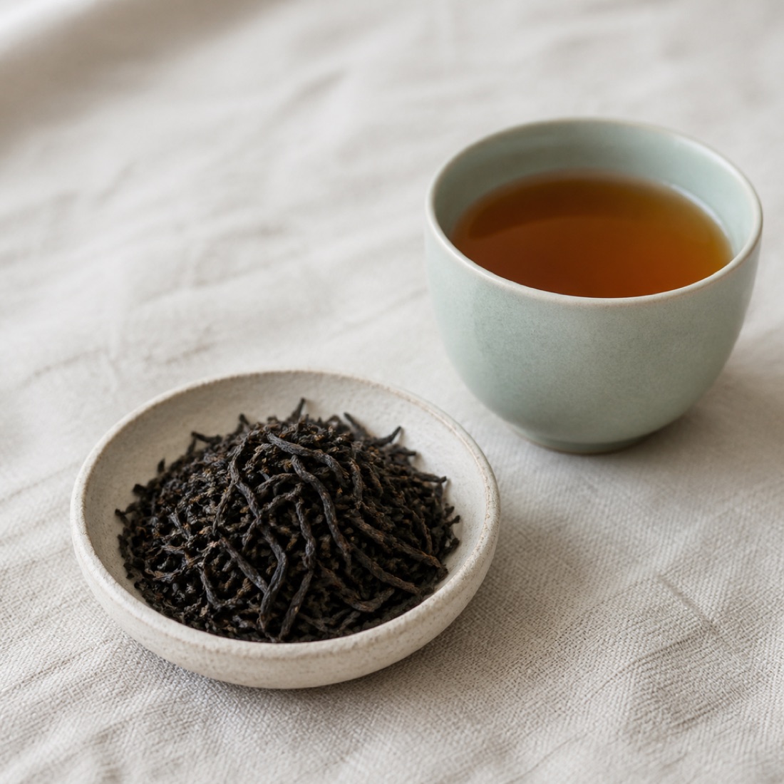 Fine wiry dark black tea leaves beside a cup of amber liquor