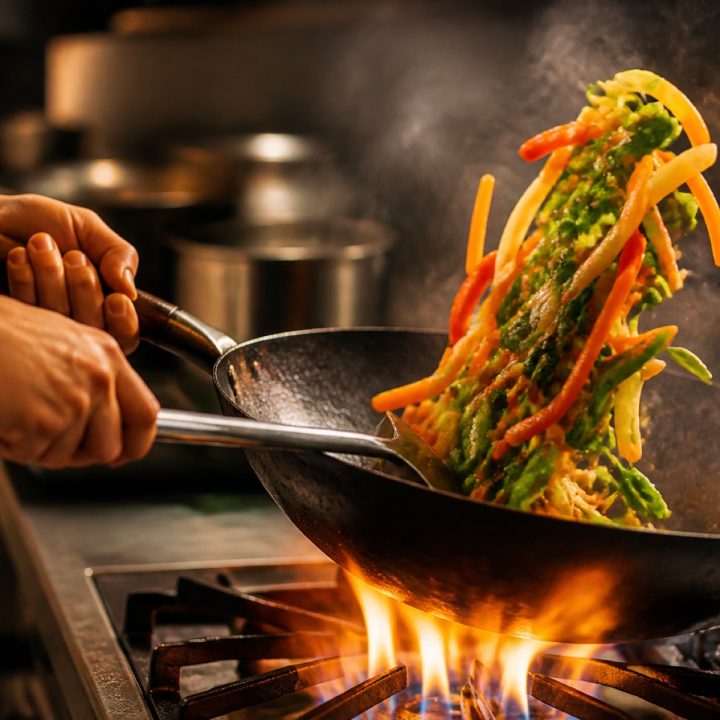 Hands tossing vegetables in a carbon steel wok over a high flame