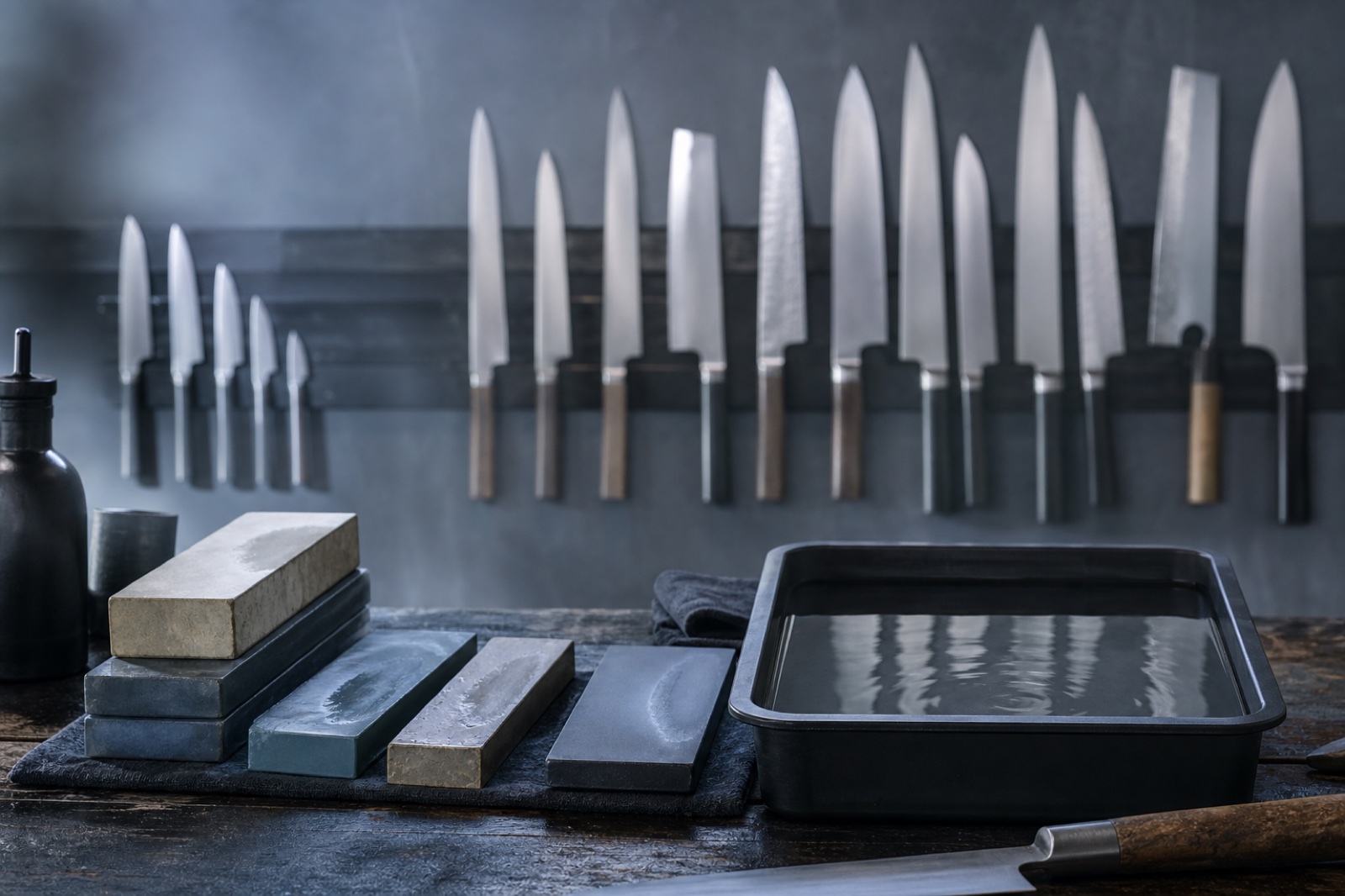 Knife sharpening workbench with whetstones and a water tray, a wall rack of kitchen knives softly blurred behind